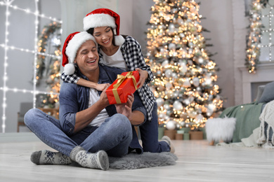 Image of Happy young couple in Santa hats with Christmas gift at home