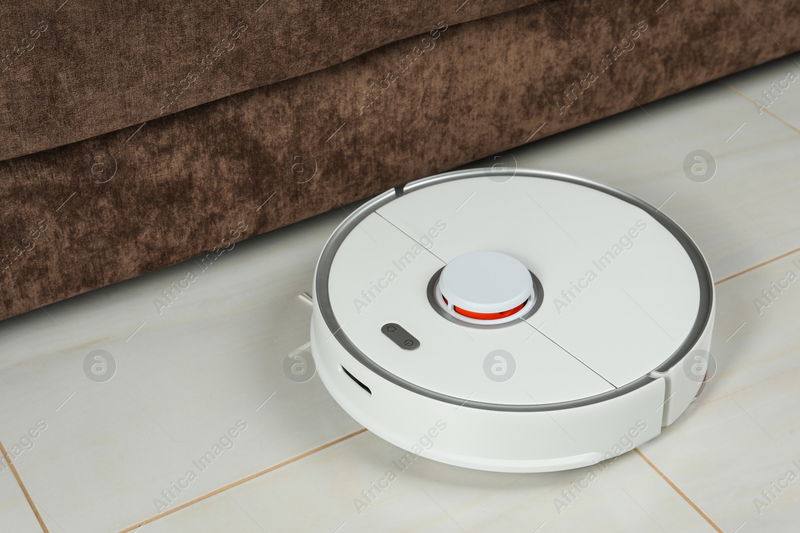 Photo of Robotic vacuum cleaner on white tiled floor near sofa indoors