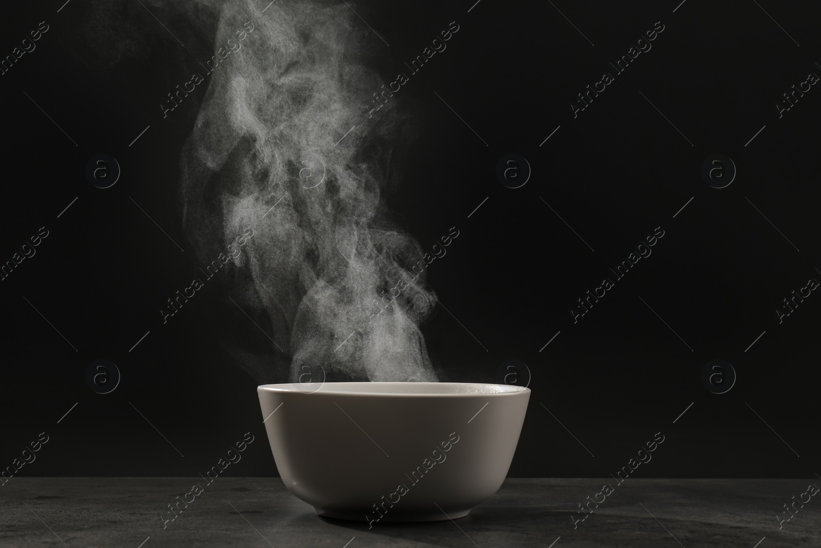 Photo of Steaming ceramic bowl on grey table against dark background