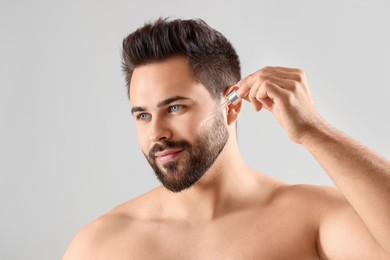 Handsome man applying cosmetic serum onto face on light grey background