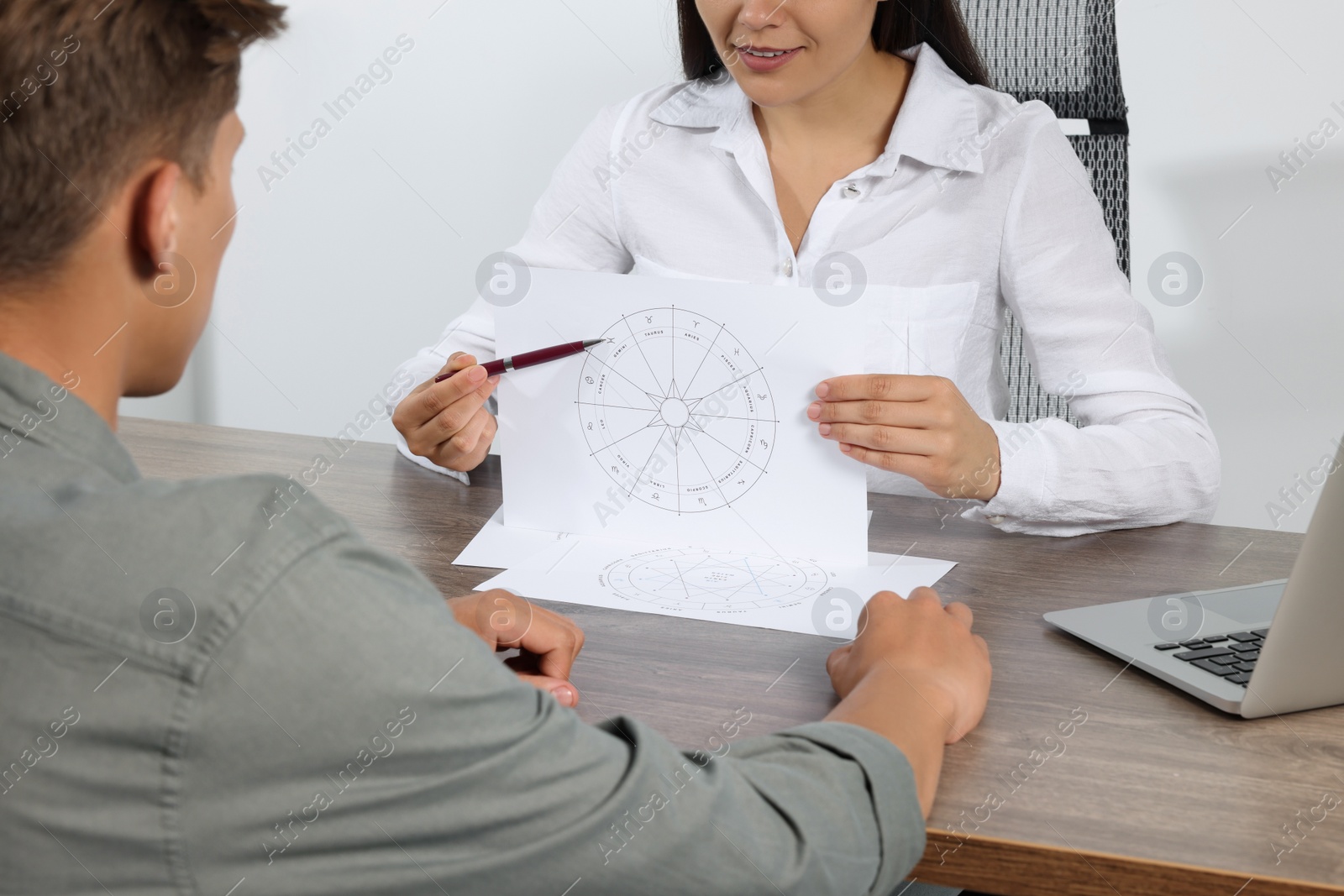 Photo of Astrologer showing zodiac wheel to client at wooden table indoors, closeup