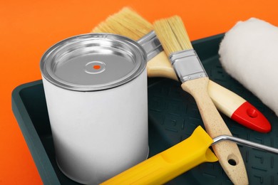 Photo of Can of orange paint, brushes, roller and container on color background, closeup