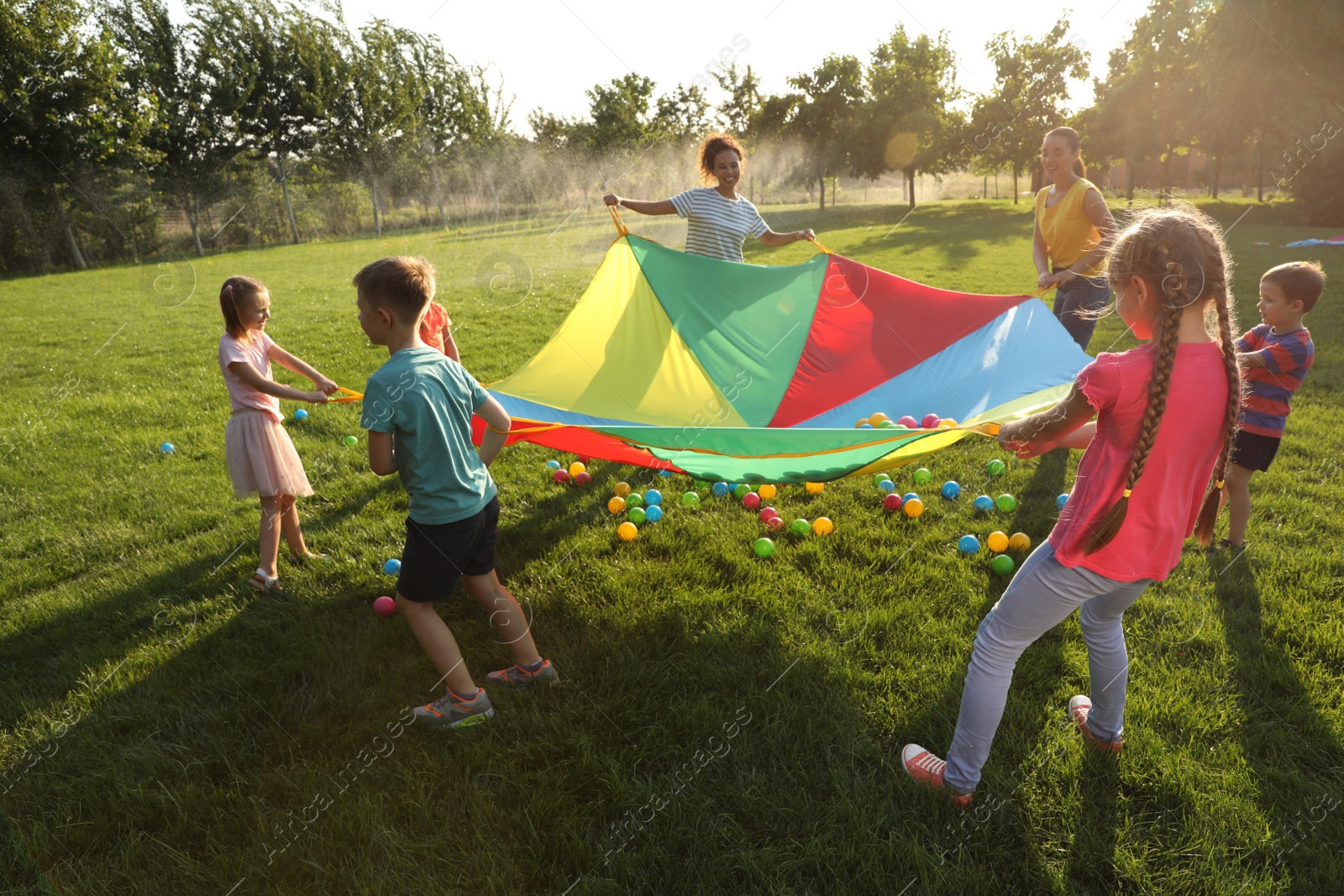 Photo of Group of children and teachers playing with rainbow playground parachute on green grass. Summer camp activity