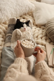 Photo of Woman holding cup of hot drink over adorable cat on knitted blanket, closeup