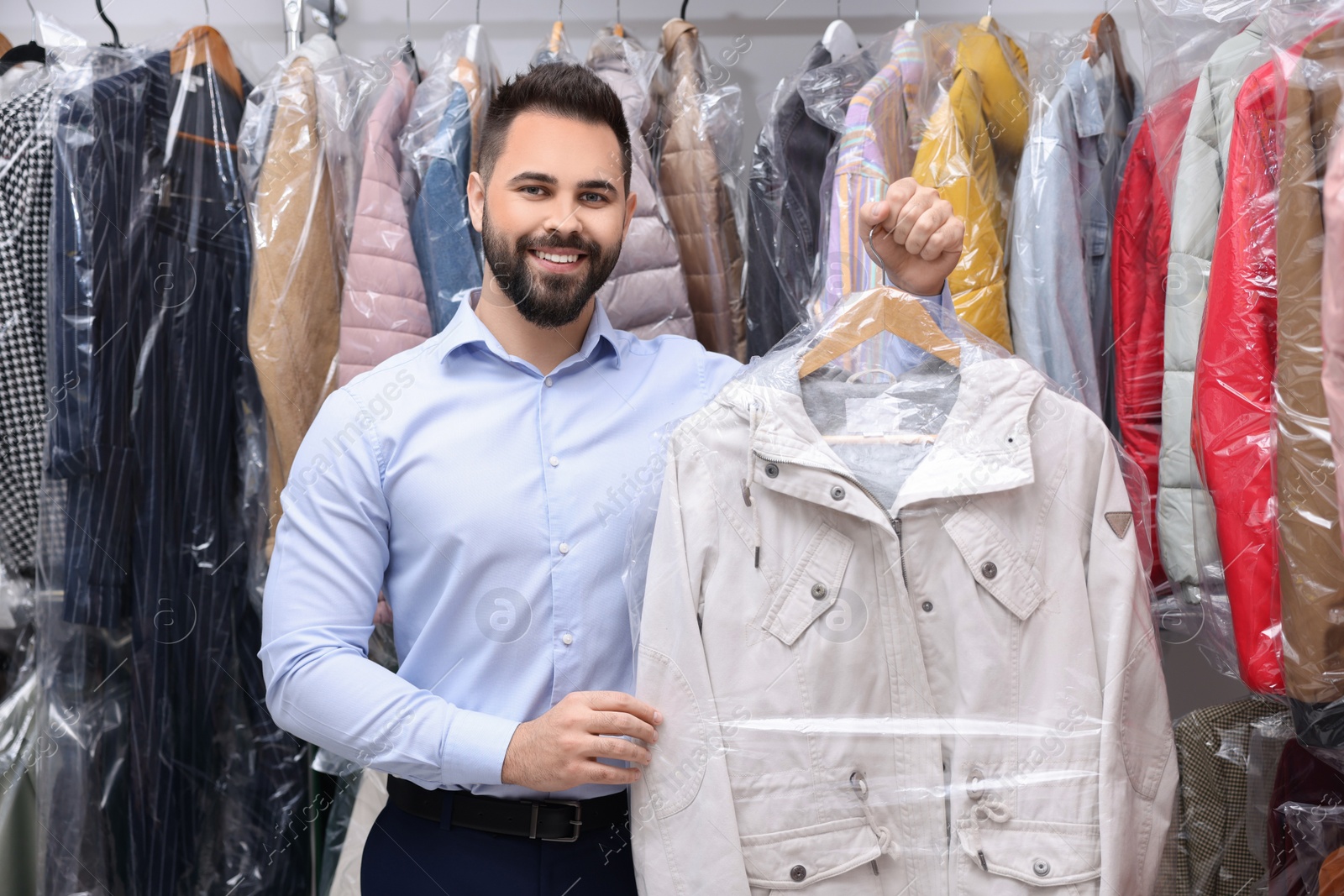 Photo of Dry-cleaning service. Happy worker holding hanger with jacket in plastic bag near other clothes indoors