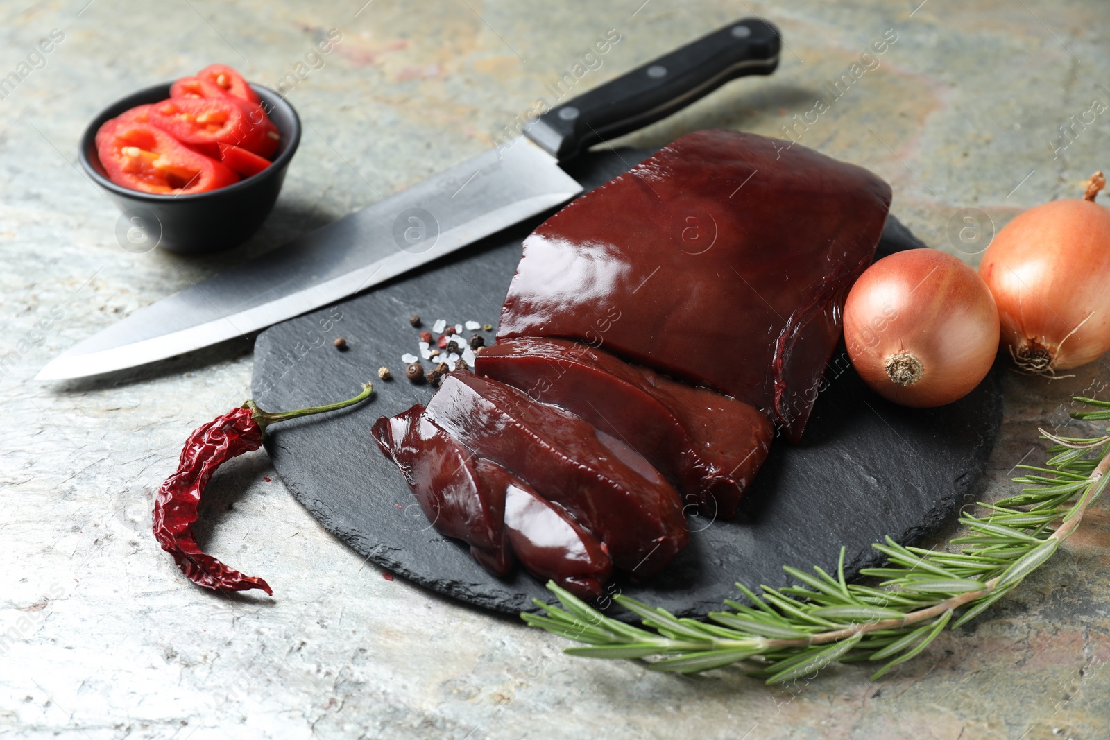 Photo of Cut raw beef liver with rosemary, chili pepper, onions and knife on grey table