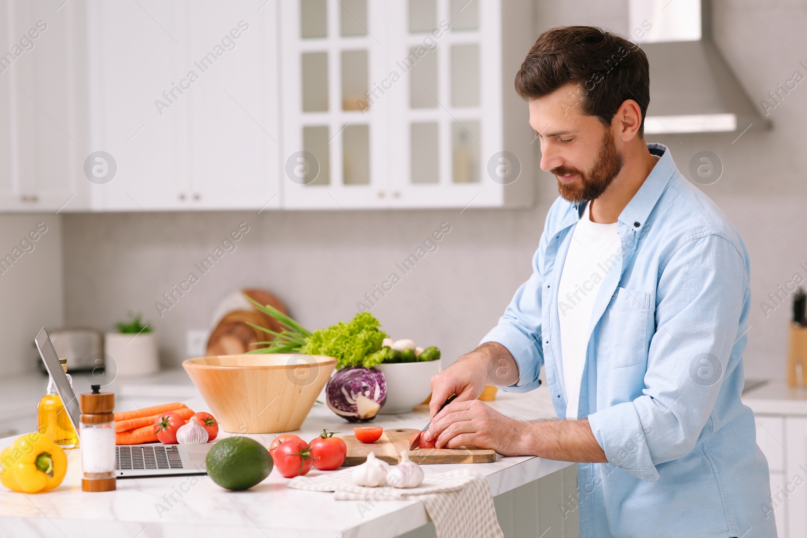 Photo of Man making dinner while watching online cooking course via laptop in kitchen