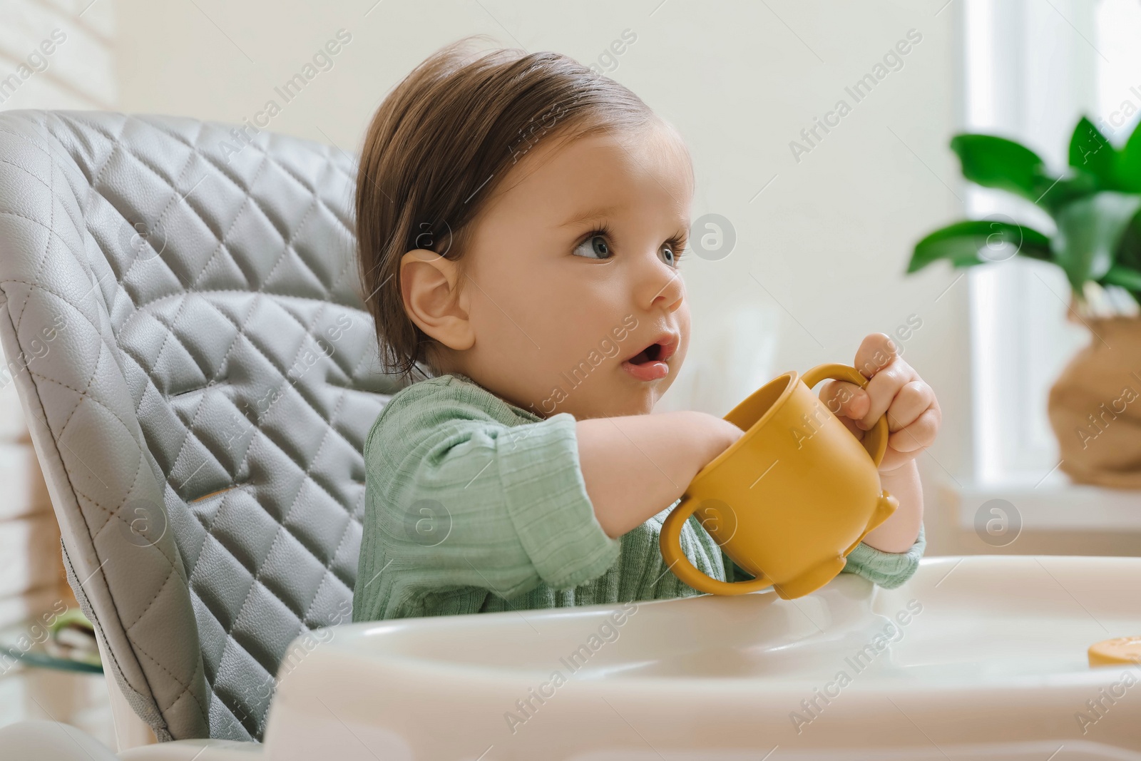 Photo of Cute little baby with cup in high chair indoors
