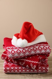 Photo of Stack of different Christmas sweaters and Santa hat on wooden table against beige background