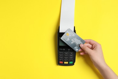 Woman using payment terminal with credit card and thermal paper for receipt on yellow background, top view. Space for text