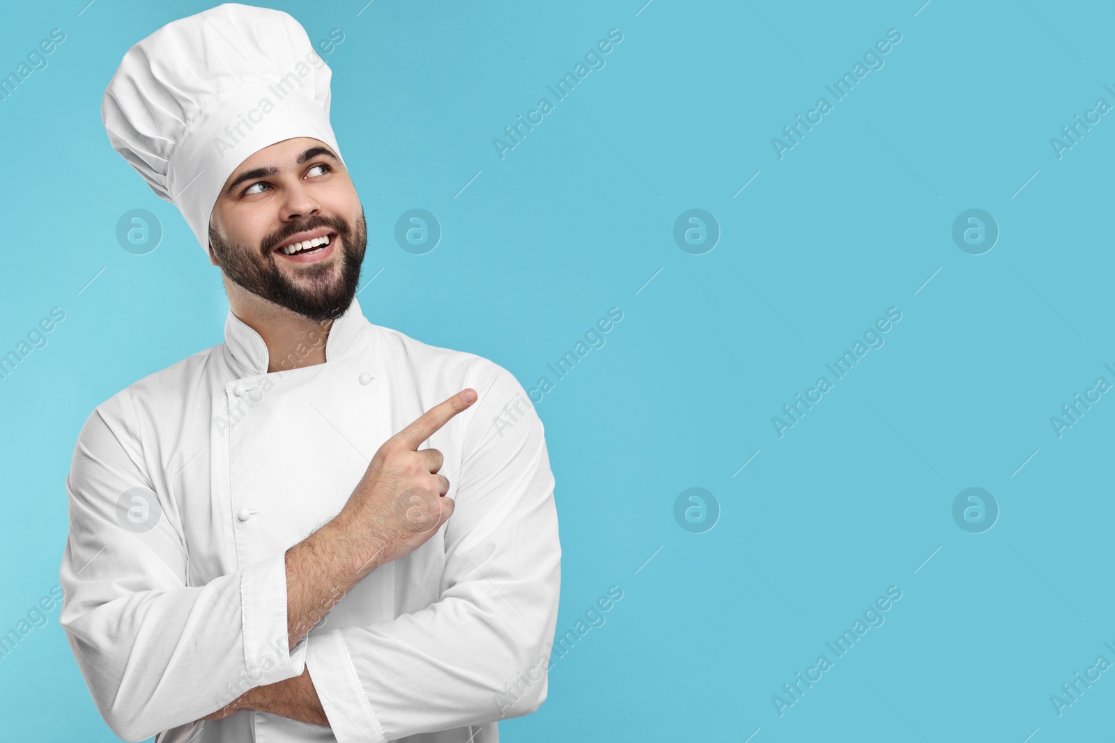 Photo of Happy young chef in uniform pointing at something on light blue background. Space for text