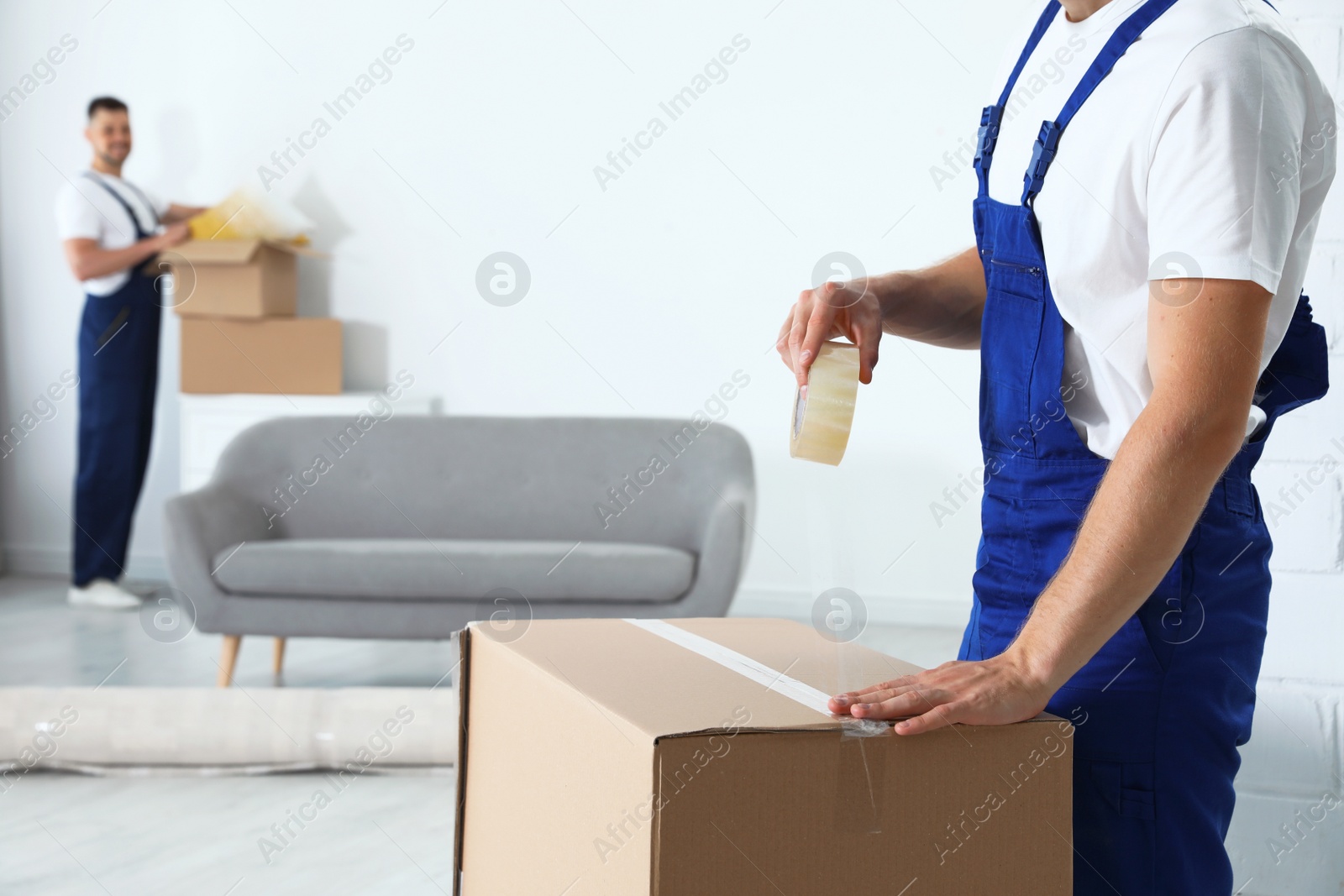 Photo of Moving service employee sealing cardboard box with adhesive tape in room, closeup