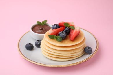 Photo of Delicious pancakes with strawberries, blueberries, mint and chocolate sauce on pink background