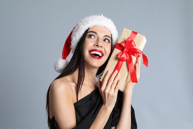 Photo of Woman in black dress and Santa hat holding Christmas gift on grey background