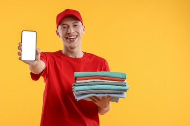 Dry-cleaning delivery. Happy courier holding folded clothes and smartphone on orange background, space for text