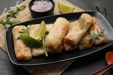 Photo of Tasty fried spring rolls, arugula, lime and sauce on dark table, closeup