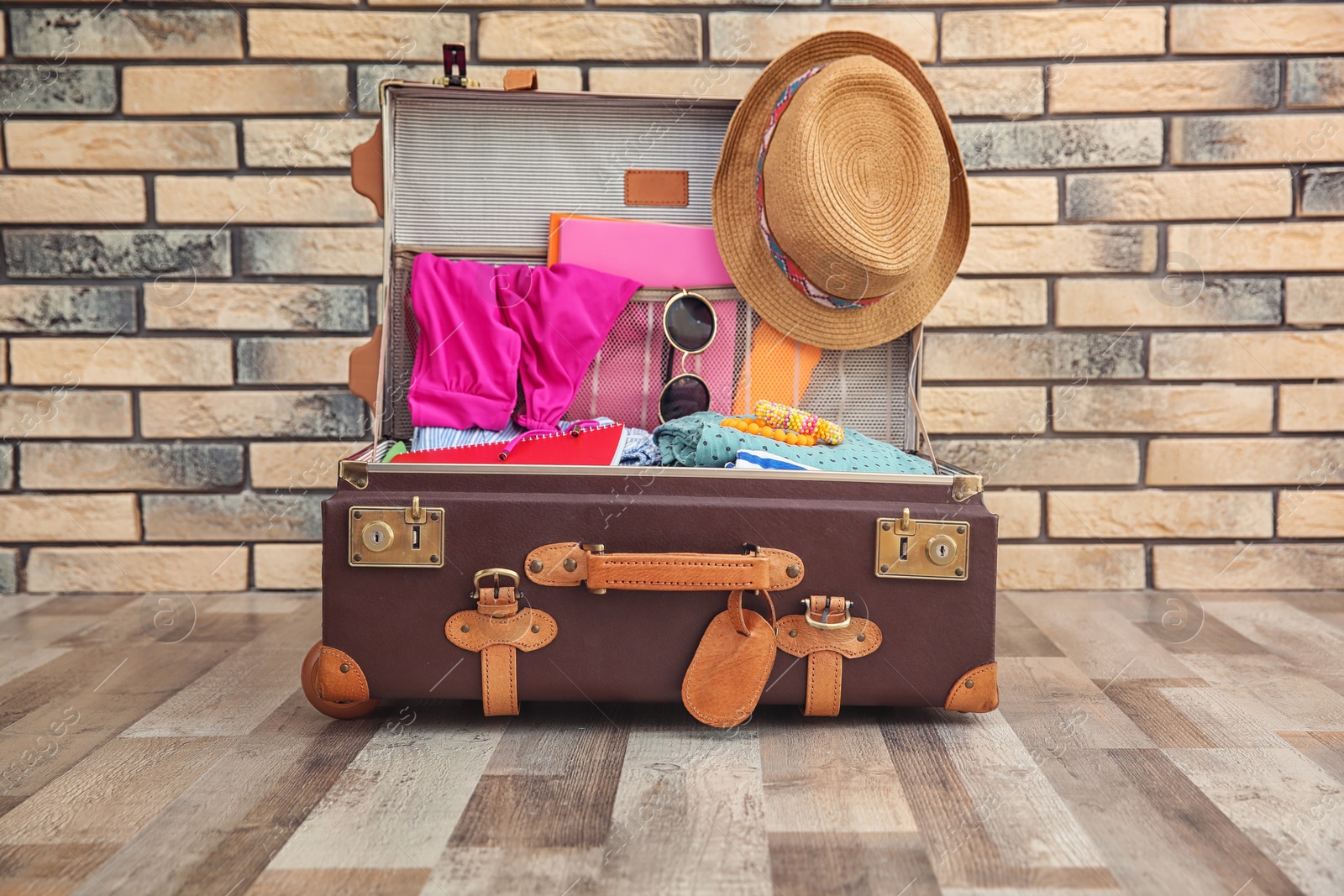 Photo of Open suitcase with different clothes and accessories for summer journey on floor near brick wall