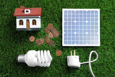 Flat lay composition with solar panel, house model, light bulb and coins on green grass