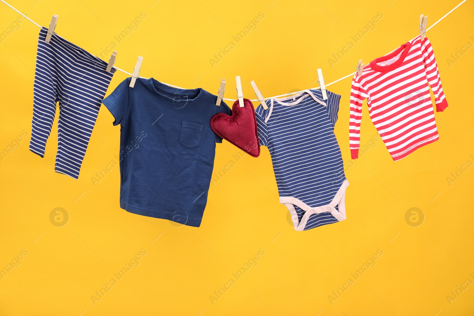 Photo of Different baby clothes and heart shaped cushion drying on laundry line against orange background