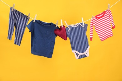 Photo of Different baby clothes and heart shaped cushion drying on laundry line against orange background