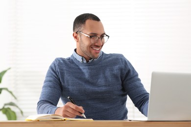 Photo of Smiling African American man writing in notebook while working on laptop at wooden table