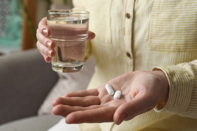 Calcium supplement. Woman holding glass of water and pills indoors, closeup