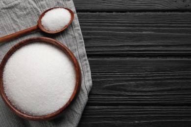 Granulated sugar in bowl and spoon on black wooden table, flat lay. Space for text