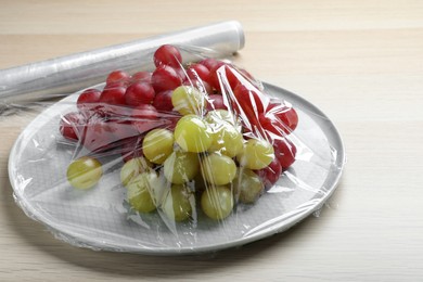 Photo of Plate of fresh grapes with plastic food wrap on wooden table, closeup