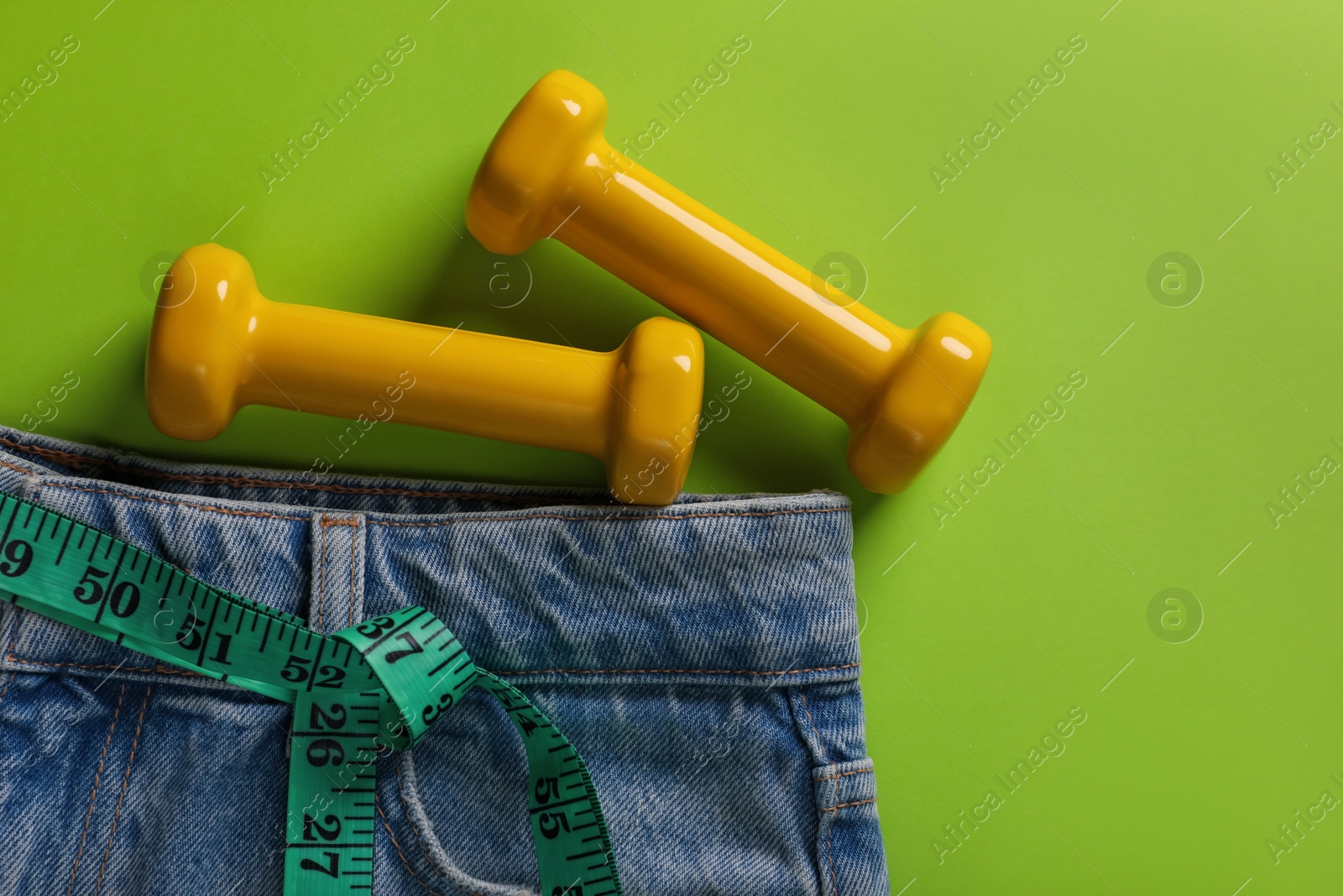 Photo of Jeans, dumbbells and measuring tape on light green background, flat lay with space for text. Weight loss concept