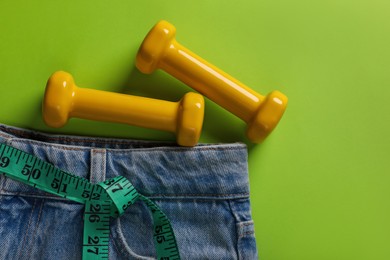 Jeans, dumbbells and measuring tape on light green background, flat lay with space for text. Weight loss concept