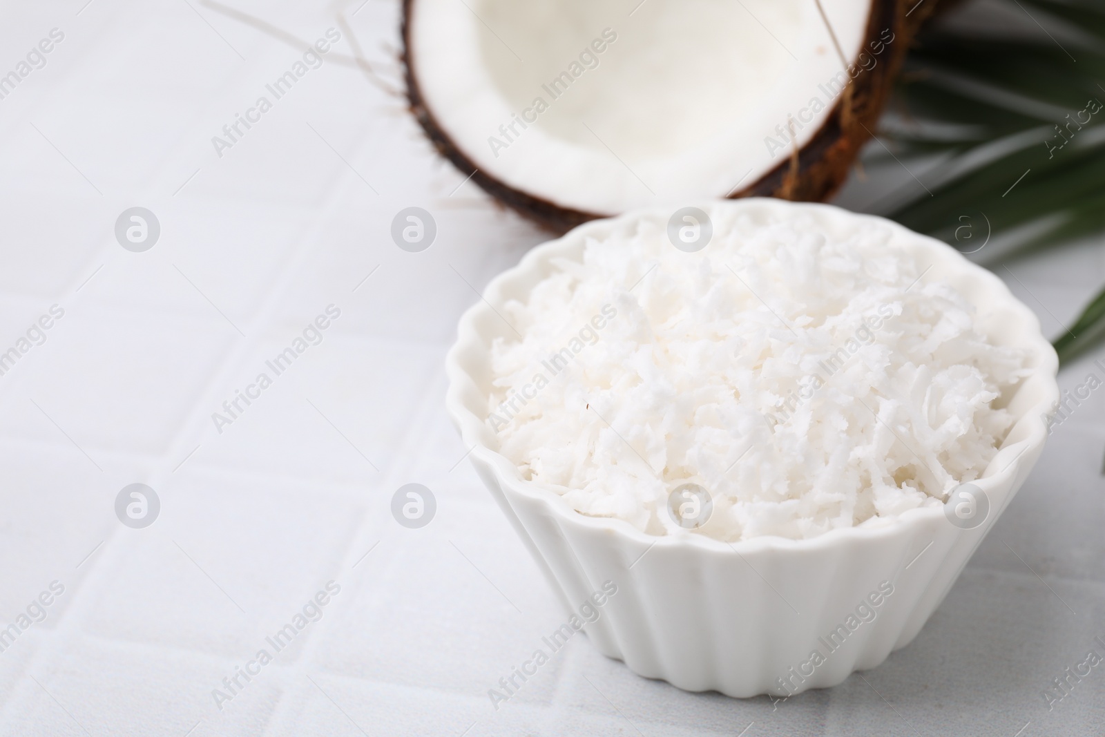 Photo of Coconut flakes in bowl, nut and palm leaf on white tiled table, closeup. Space for text