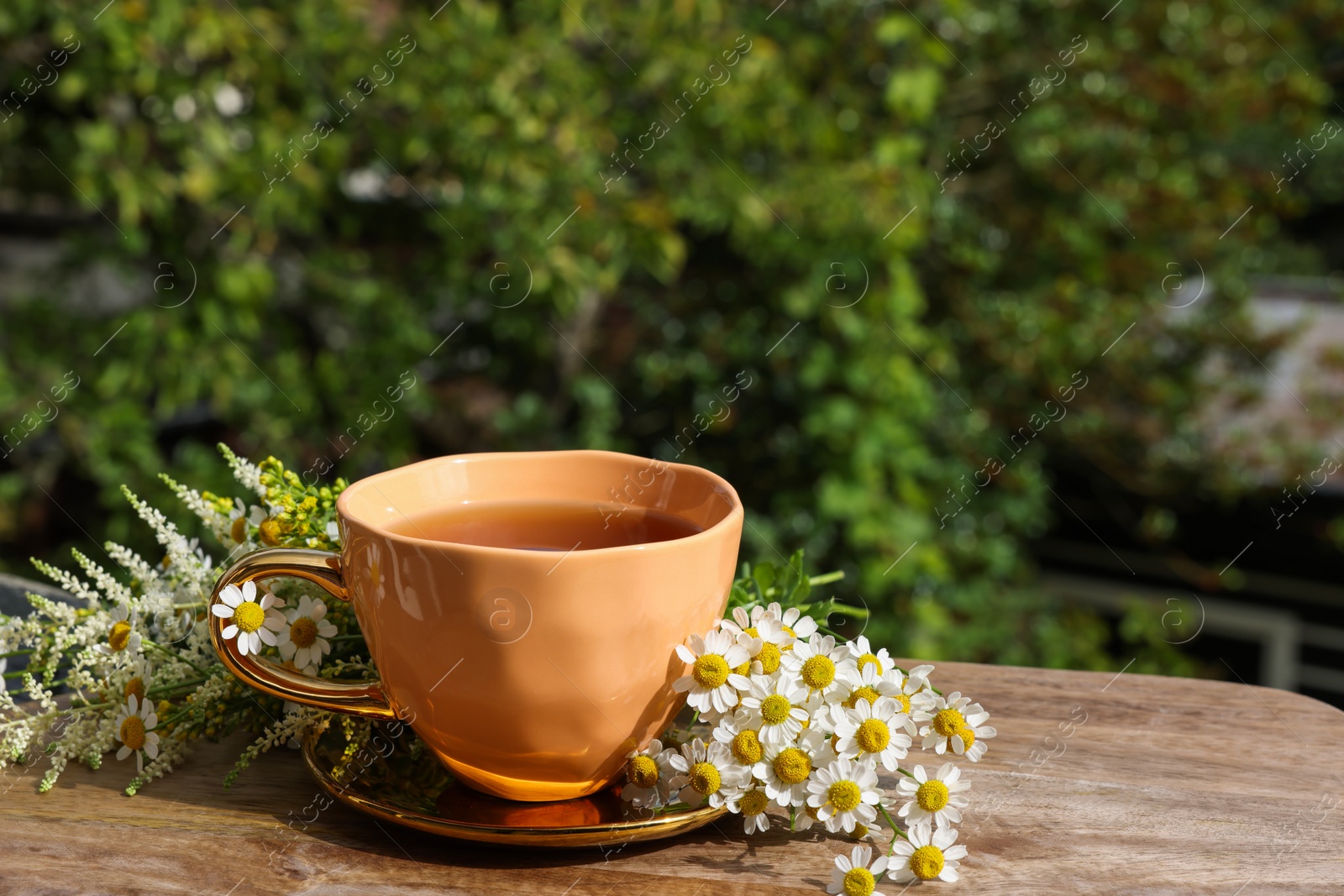 Photo of Cup of delicious chamomile tea and fresh flowers outdoors on sunny day, space for text