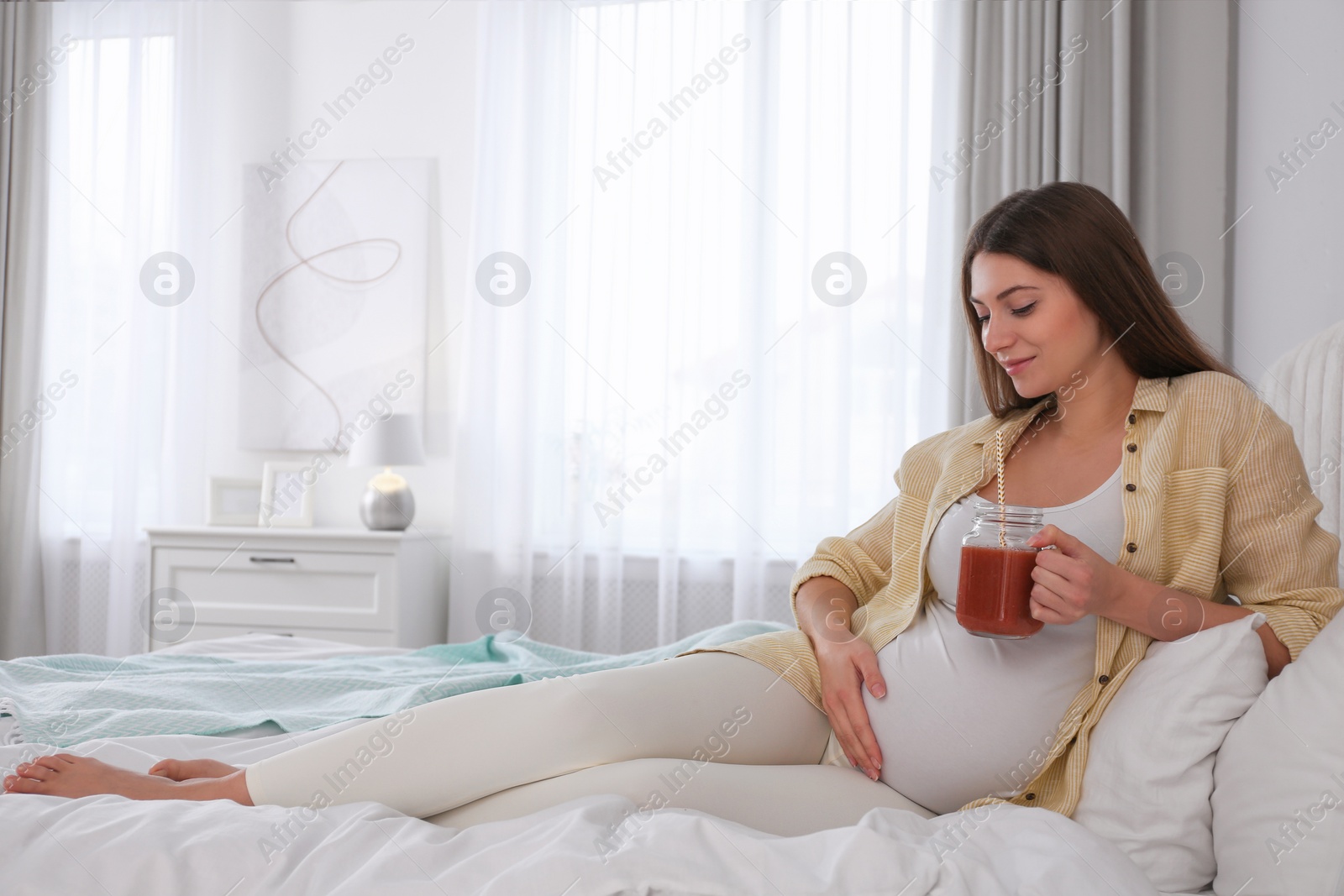 Photo of Pregnant woman with smoothie on bed at home. Healthy diet