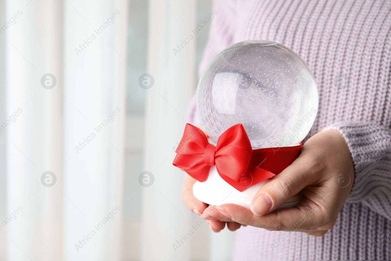 Photo of Woman holding empty snow globe with red bow indoors, closeup. Space for text