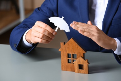 Man covering wooden houses with umbrella cutout at table, closeup. Home insurance