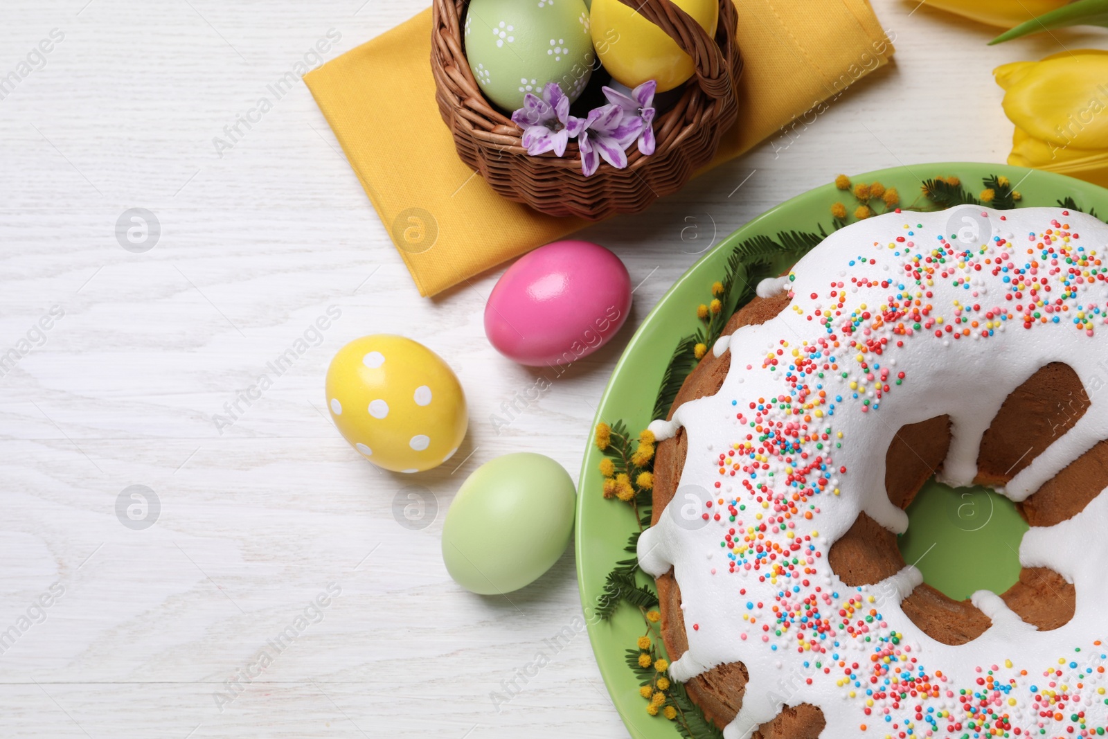 Photo of Glazed Easter cake with sprinkles, painted eggs and flowers on white wooden table, flat lay. Space for text