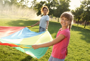 Cute child and African-American teacher playing with rainbow playground parachute on green grass. Summer camp activity