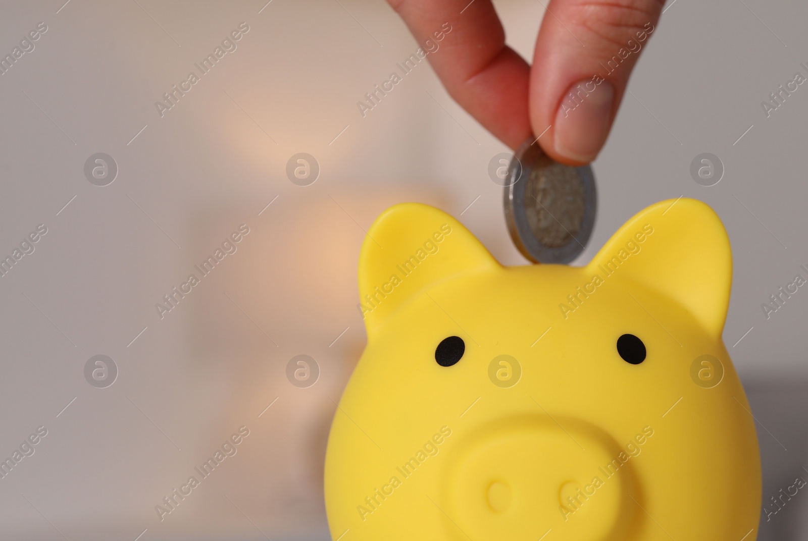 Photo of Woman putting coin into yellow piggy bank on blurred background, closeup