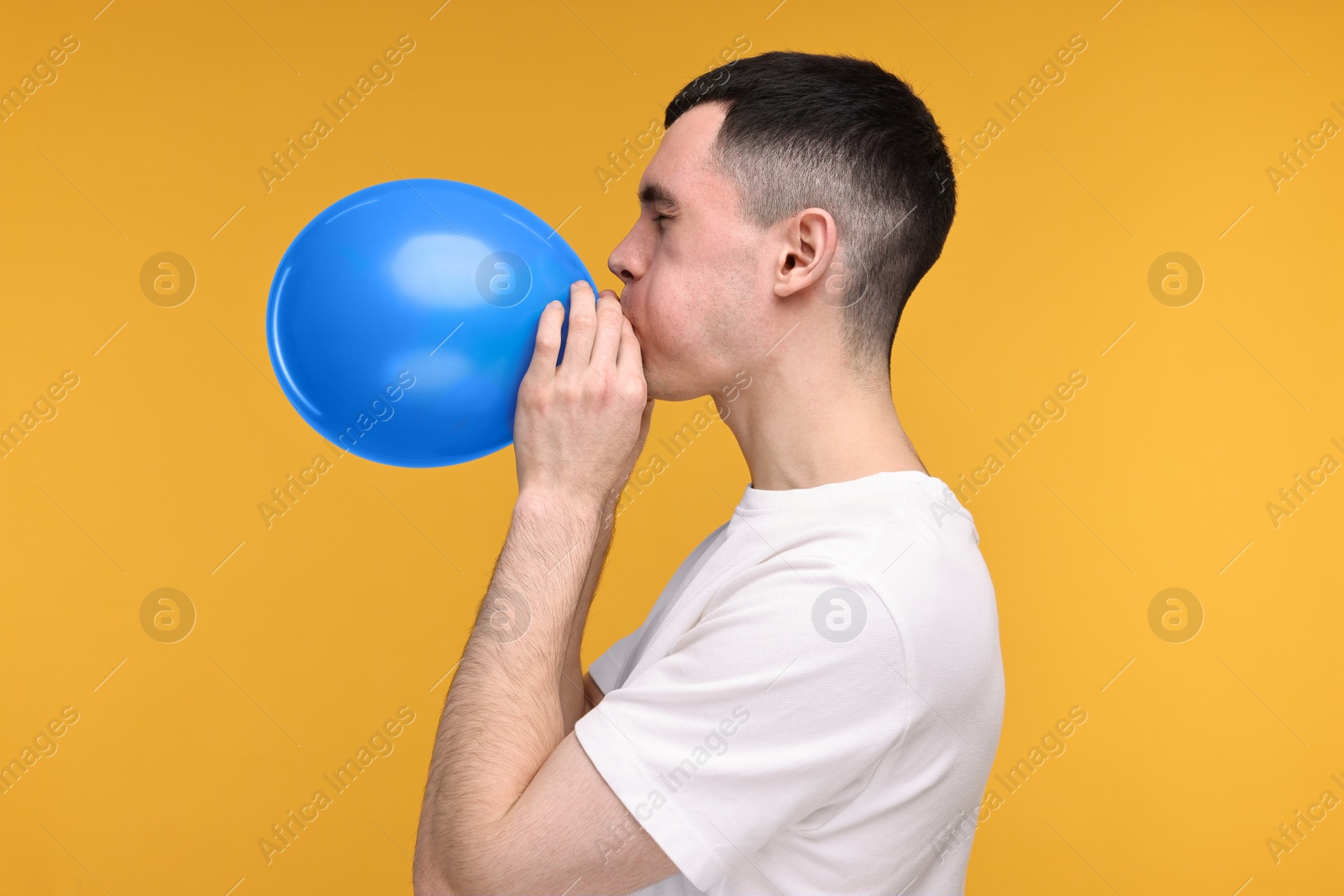 Photo of Young man inflating light blue balloon on yellow background