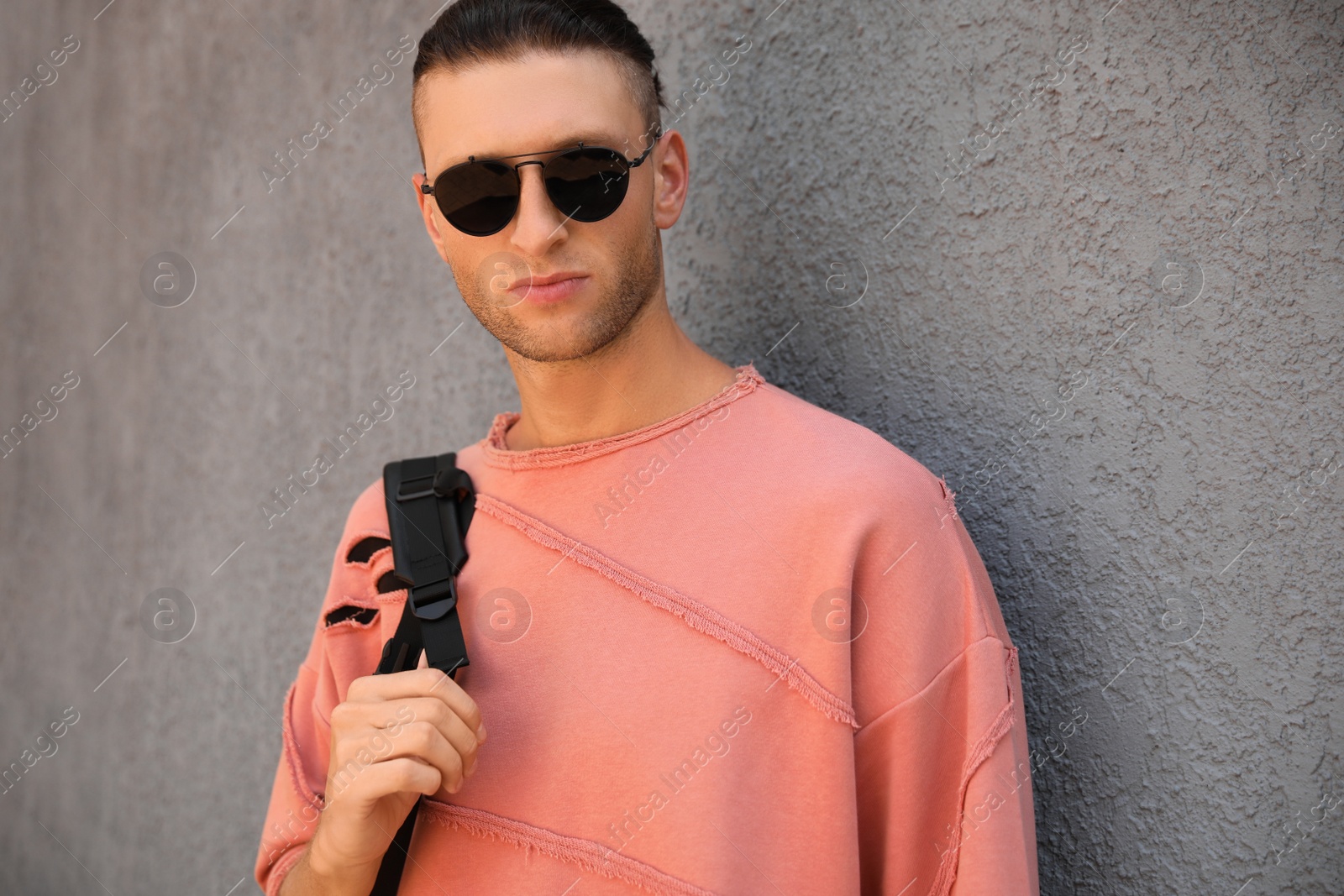 Photo of Handsome young man in stylish sunglasses and backpack near grey wall