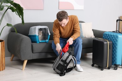 Photo of Travel with pet. Man putting headphones into backpack near carrier with cute cat at home