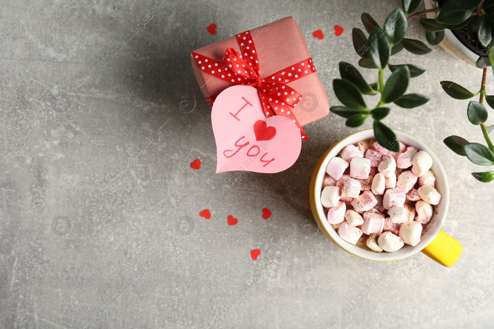 Photo of Heart shaped greeting card with phrase I Love You attached to gift box near cup and houseplant on light grey table, flat lay. Space for text