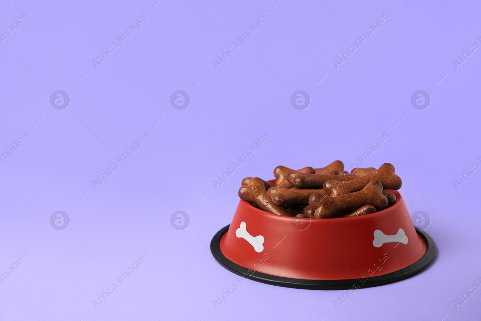 Photo of Red bowl with bone shaped dog cookies on purple background, space for text