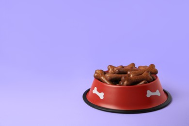 Red bowl with bone shaped dog cookies on purple background, space for text