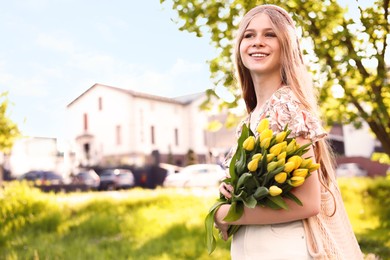 Beautiful teenage girl with bouquet of yellow tulips in city park on sunny day, space for text