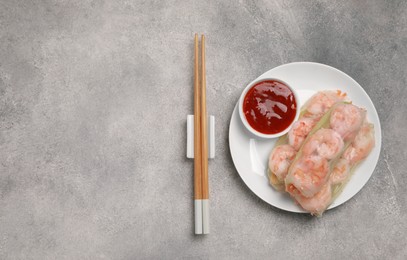 Photo of Tasty spring rolls with sauce and chopsticks on grey textured table, flat lay. Space for text