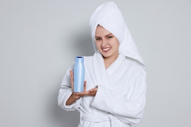 Beautiful young woman in bathrobe holding bottle of shampoo on light grey background