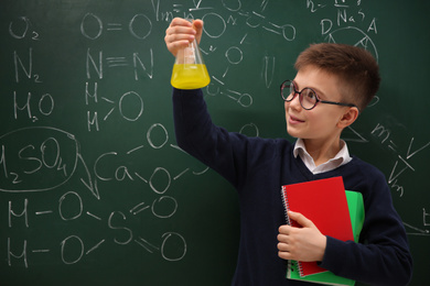 Photo of Schoolboy holding flask and notebooks near chalkboard with chemical formulas