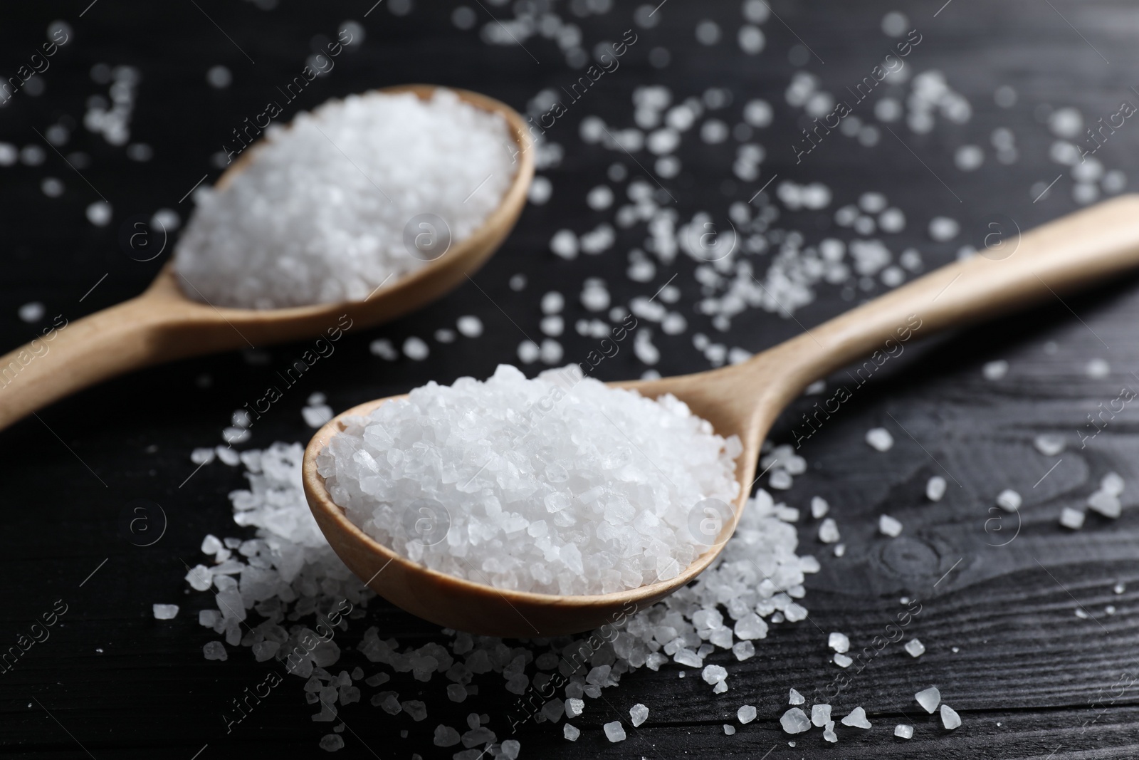 Photo of Organic salt in spoons on black wooden table, closeup