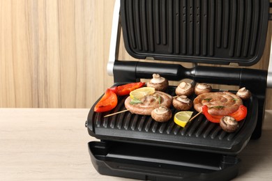 Electric grill with homemade sausages, mushrooms and bell pepper on wooden table, space for text